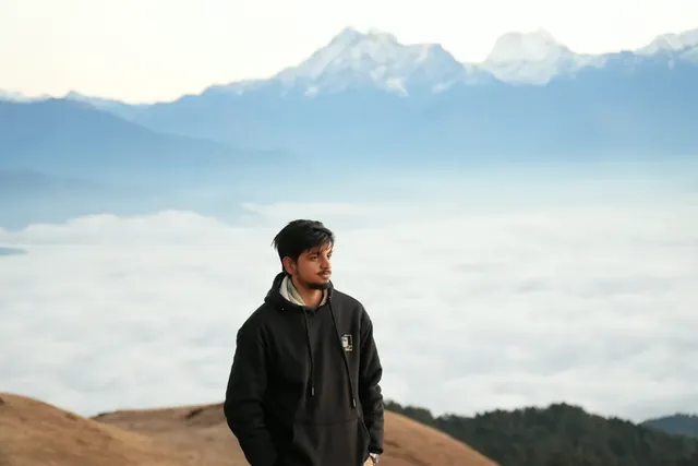Anuj Paudel in a black hoodie on a grassy Sailung ridge, with a sea of clouds and Himalayan snow peaks behind him