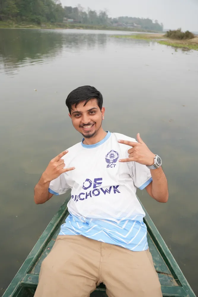Anuj Paudel smiling in an IOE Pulchowk jersey, making hand signs from a green wooden boat on calm water, Kathmandu