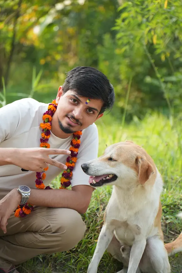 Anuj Paudel in a marigold and makhamali garland and a tika, crouching beside a tan and white dog during Tihar, Nepal