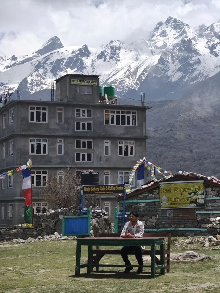 A person sits at a green wooden table outside a bakery cafe, with snow-covered peaks behind, Kyanjin Gompa