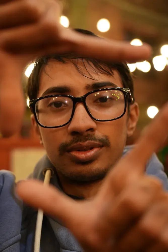 A person in glasses makes a finger frame around their face as warm string lights glow behind, campus Christmas