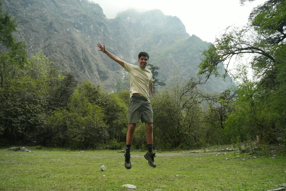 A hiker in a pale yellow T-shirt leaps into the air on a grassy clearing below a steep, misty valley wall in Langtang