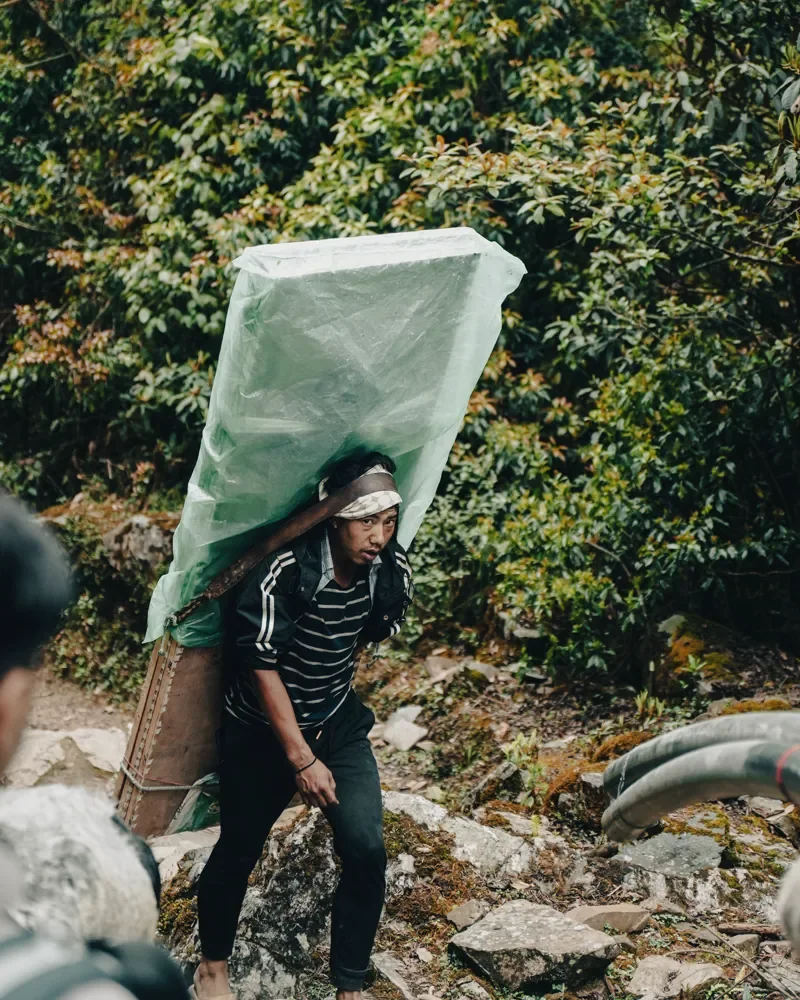 A porter in a striped top carries a large load wrapped in green plastic along a rocky forest trail at Panch Pokhari