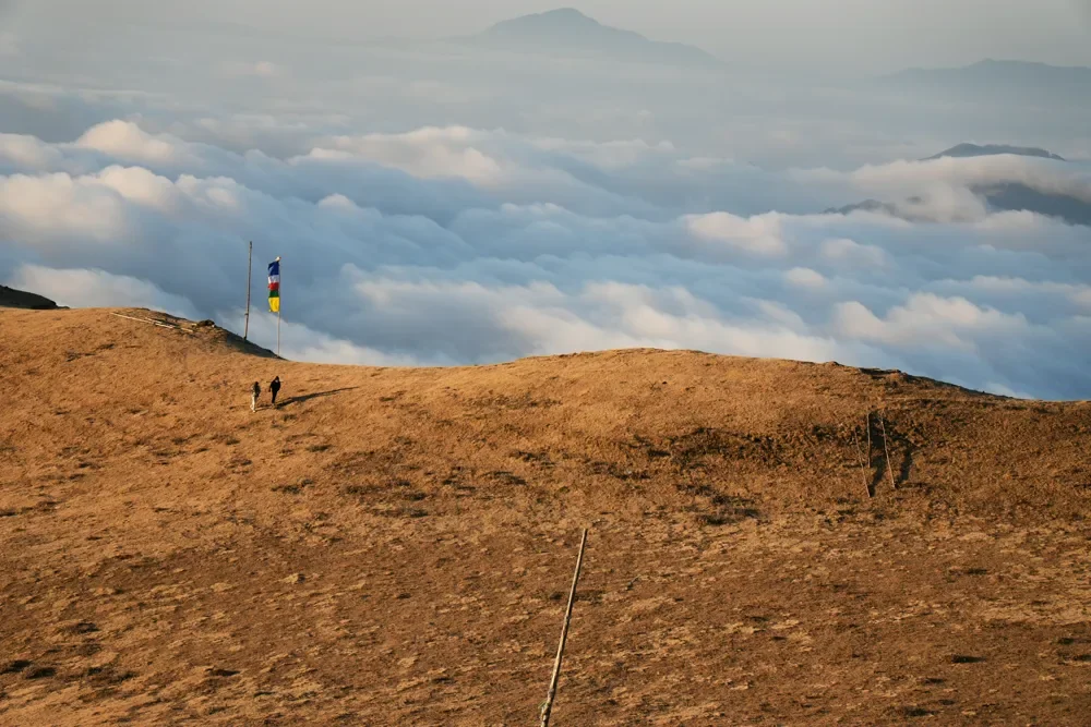 Two trekkers on a golden grass ridge below a prayer flag, with a sea of clouds beyond, Sailung