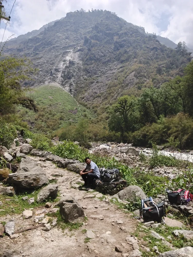 A man sits on a rock by a rocky trail, small against a huge forested ridge above a river in the Langtang valley