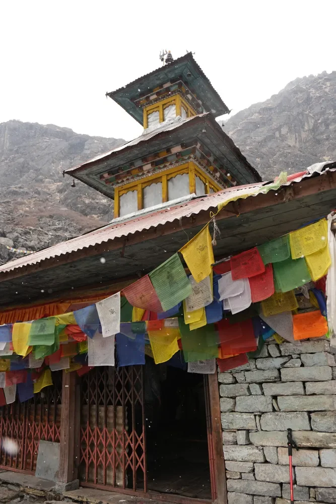 Two-tiered temple with a red lattice gate and rows of prayer flags under light snowfall, Panch Pokhari