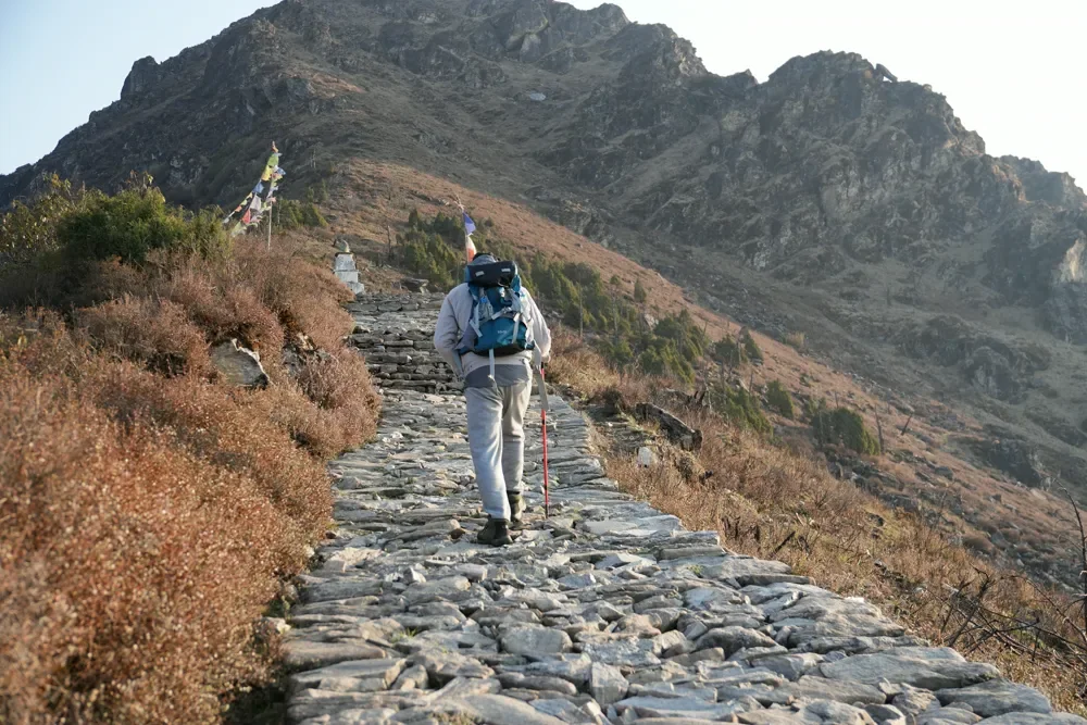 A trekker with a blue backpack walks up a stone-paved path toward a rocky ridge and prayer flags at Panch Pokhari