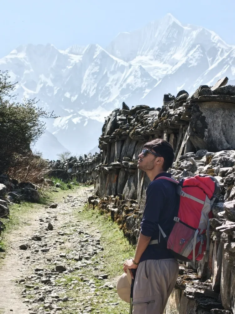 A trekker in sunglasses and a red backpack looks up at snow peaks beside a long stone wall in the Langtang valley