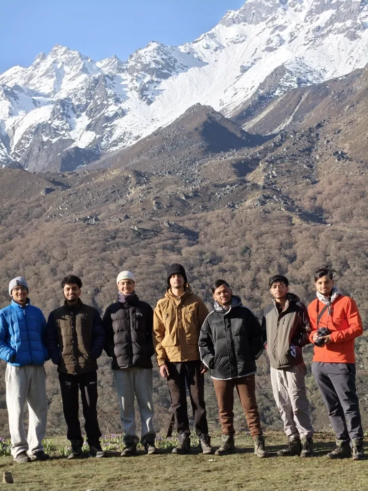 Seven trekkers in puffer jackets stand in a row on a grassy slope with snow-covered peaks behind them in Langtang