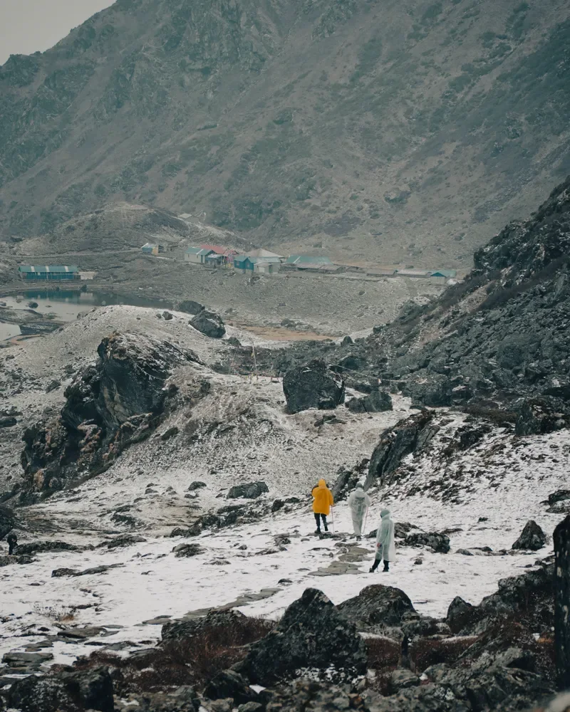 Trekkers in rain ponchos walk along a snowy path toward a small lake village at Panch Pokhari