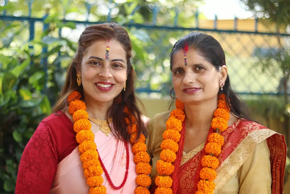 Two smiling women in orange marigold garlands and bright tika marks, standing outdoors during Bhai Tika